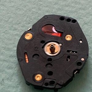 May include: A black watch movement with a red component and gold-coloured accents. The movement features a small gear in the centre and several holes and circular components. The background is a light green colour.