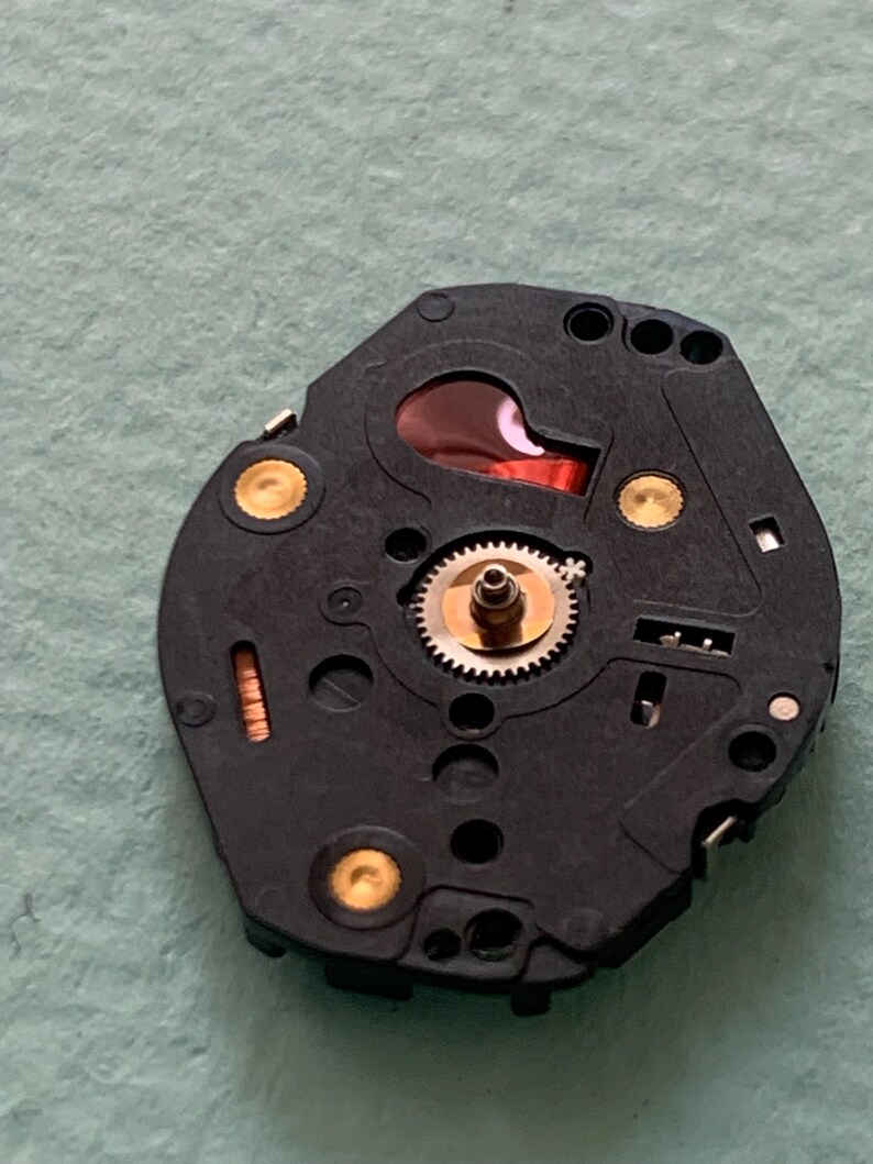 May include: A black watch movement with a red component and gold-coloured accents. The movement features a small gear in the centre and several holes and circular components. The background is a light green colour.