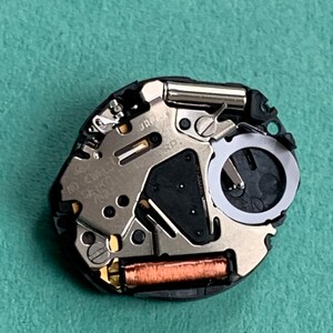 May include: Close-up of a watch movement with intricate metal components, including a silver-colored plate, a black section, and a copper-colored coil. The watch mechanism is set against a green background.