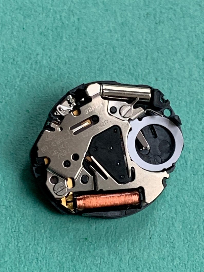 May include: Close-up of a watch movement with intricate metal components, including a silver-colored plate, a black section, and a copper-colored coil. The watch mechanism is set against a green background.