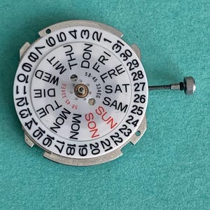 May include: A white watch calendar dial with black numbers and day abbreviations. The day of the week is printed in black, with "SUN" in red. A small screw extends from the side. The dial has the numbers 1-31.