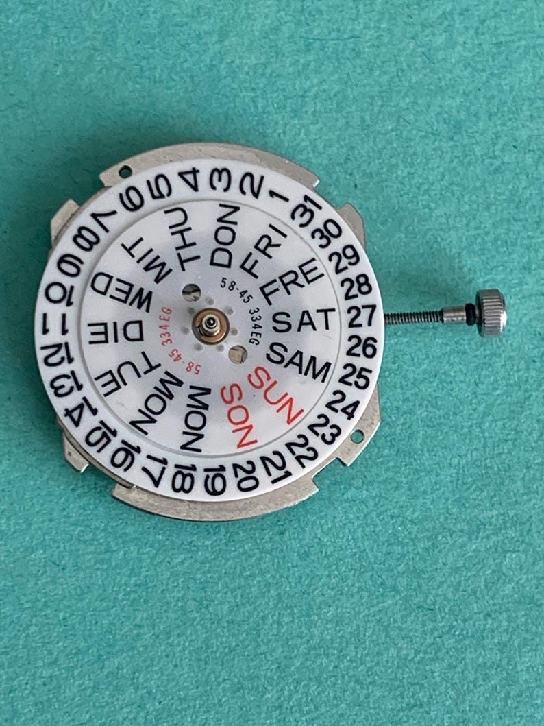 May include: A white watch calendar dial with black numbers and day abbreviations. The day of the week is printed in black, with "SUN" in red. A small screw extends from the side. The dial has the numbers 1-31.
