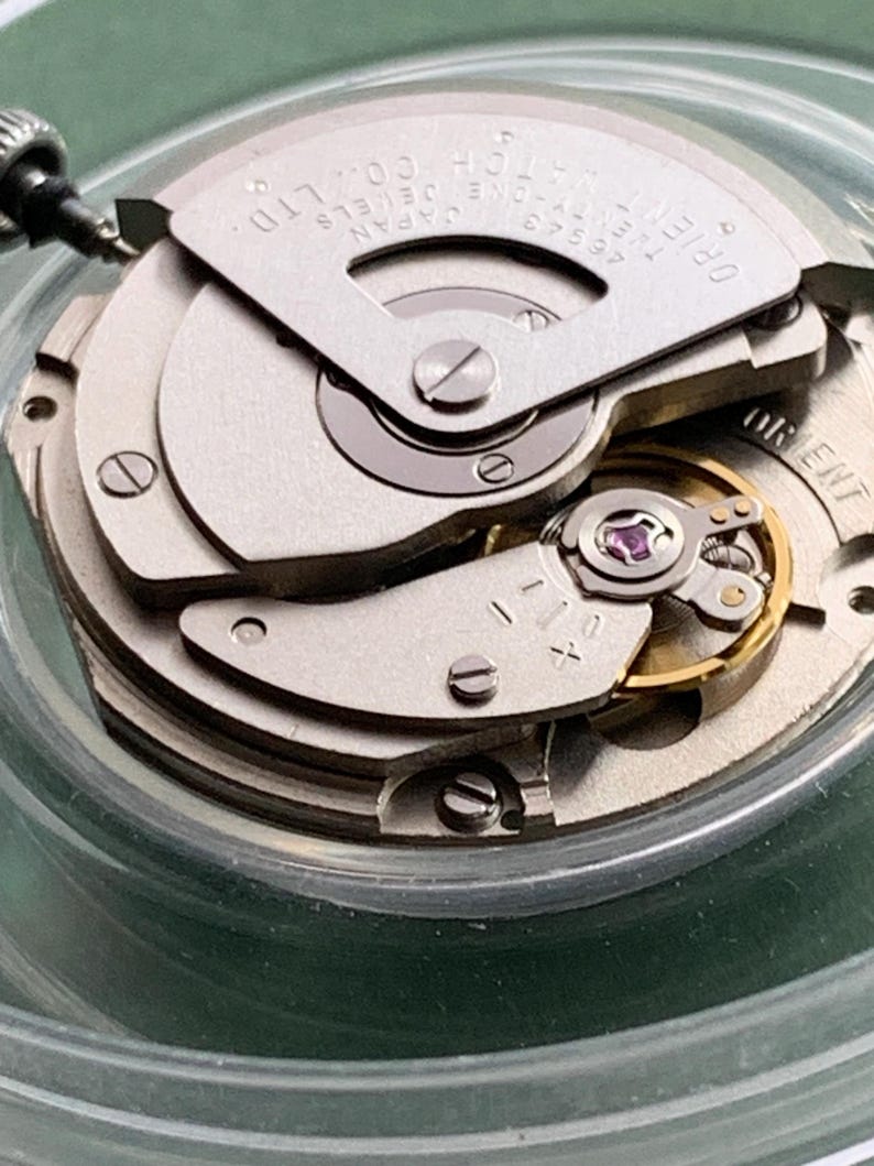 May include: Detailed view of a watch movement with silver and gold parts. The mechanism displays intricate gears, screws, and a balance wheel. The text "ORIENT WATCH CO., LTD." is visible on a silver plate. The watch is set in a clear, round container.