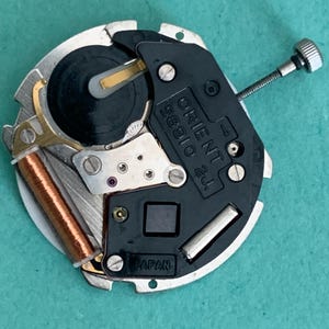 May include: A close-up of a watch movement, featuring the text "ORIENT 58910 2J" and "JAPAN". The mechanism has silver, black, and copper parts, including a small screw and a coil. The watch movement is circular in shape.