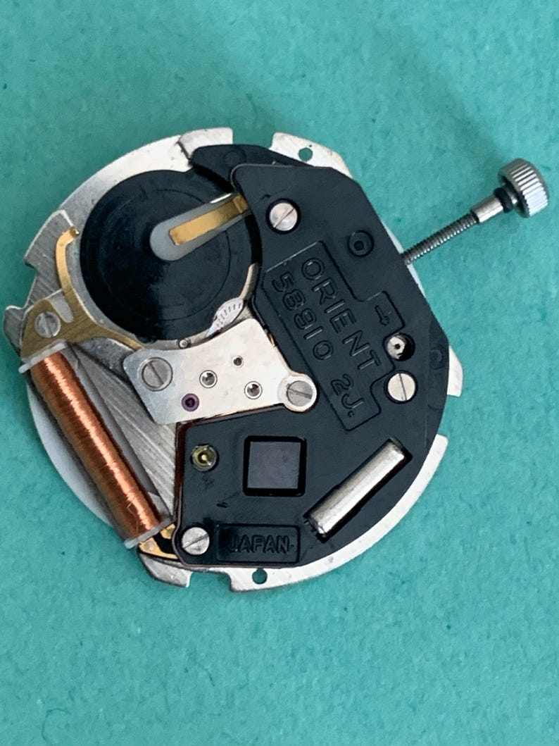 May include: A close-up of a watch movement, featuring the text "ORIENT 58910 2J" and "JAPAN". The mechanism has silver, black, and copper parts, including a small screw and a coil. The watch movement is circular in shape.