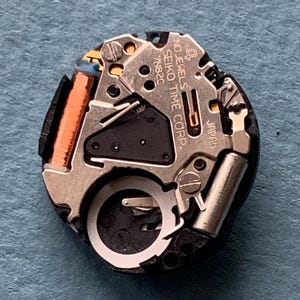 K&ouml;nnte beinhalten: Nahaufnahme eines Seiko-Uhrwerks. Der Mechanismus umfasst silberfarbene Metallkomponenten, eine kupferfarbene Spule und den Text "SEIKO TIME CORP" und "JAPAN". Das Uhrwerk befindet sich vor einem blauen Hintergrund.