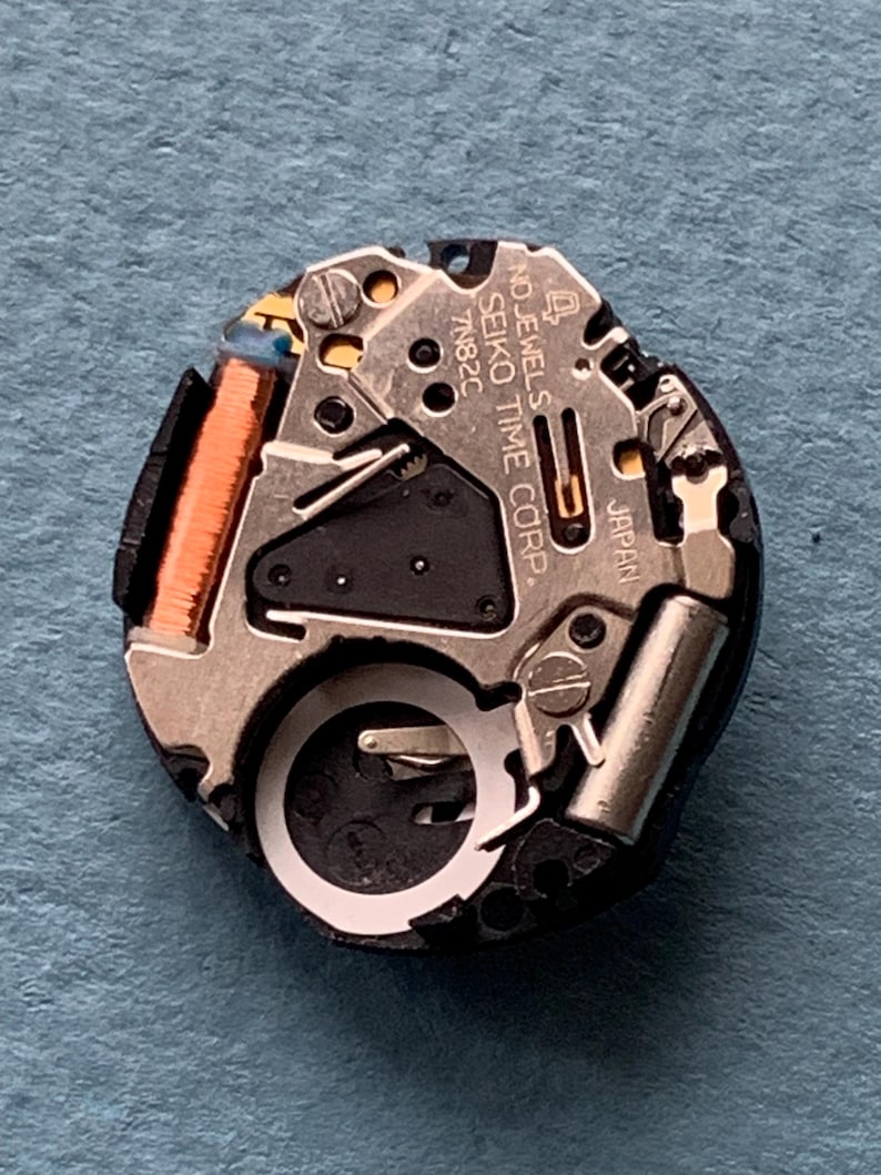 K&ouml;nnte beinhalten: Nahaufnahme eines Seiko-Uhrwerks. Der Mechanismus umfasst silberfarbene Metallkomponenten, eine kupferfarbene Spule und den Text "SEIKO TIME CORP" und "JAPAN". Das Uhrwerk befindet sich vor einem blauen Hintergrund.