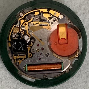 May include: Close-up of a Citizen watch movement. The watch mechanism features gold-colored gears and components, a red battery, and the text "CITIZEN WATCH CO". The watch case is dark green, and the crown is silver.