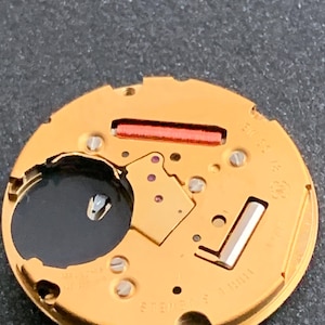 May include: Close-up of a gold-colored watch movement with various components, including screws, a red rectangular piece, and a black circular element. The watch mechanism has the text "SWISS" and "3 JEWELS" printed on it.
