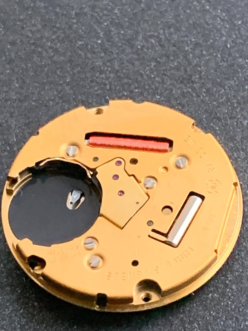 May include: Close-up of a gold-colored watch movement with various components, including screws, a red rectangular piece, and a black circular element. The watch mechanism has the text "SWISS" and "3 JEWELS" printed on it.