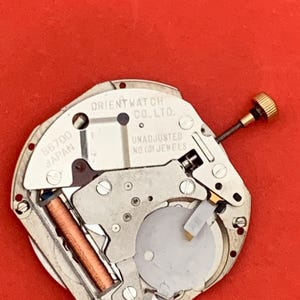 K&ouml;nnte beinhalten: Nahaufnahme eines Uhrwerks mit der Aufschrift "ORIENT WATCH CO., LTD." und "UNADJUSTED NO (0) JEWELS." Das Uhrwerk weist verschiedene Metallkomponenten, eine goldfarbene Krone und eine kupferfarbene Spule auf, vor rotem Hintergrund.