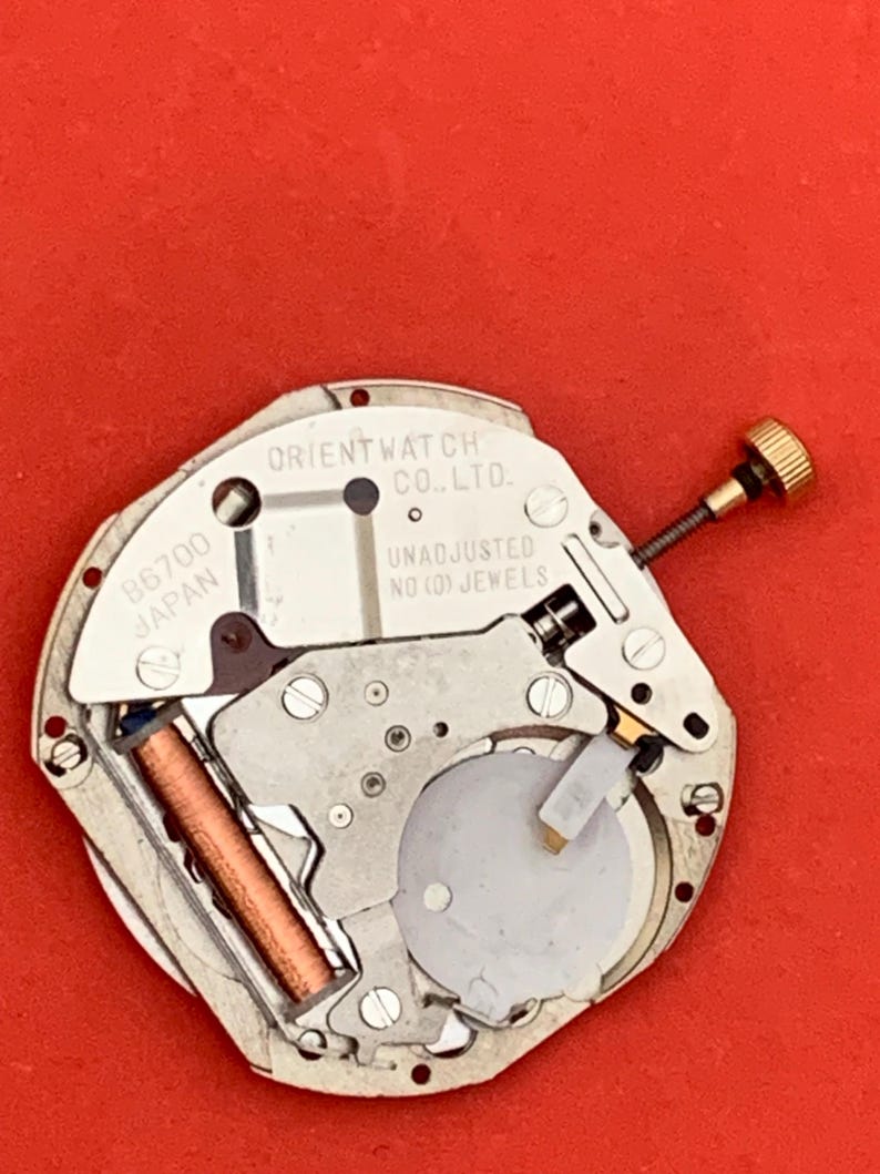 K&ouml;nnte beinhalten: Nahaufnahme eines Uhrwerks mit der Aufschrift "ORIENT WATCH CO., LTD." und "UNADJUSTED NO (0) JEWELS." Das Uhrwerk weist verschiedene Metallkomponenten, eine goldfarbene Krone und eine kupferfarbene Spule auf, vor rotem Hintergrund.