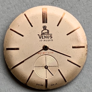 May include: A white watch face with gold-coloured markings and hands. The watch face has the text "VENUS 17 RUBIS" and "ANTIMAGNETIC" printed on it. The watch face also has the text "SWISS MADE" printed on it.