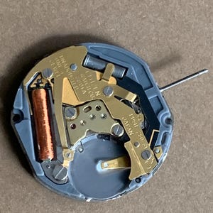 May include: A close-up of a watch movement with gold-colored gears, a copper-colored coil, and a gray plastic base. The text "CITIZEN WATCH CO." and "JAPAN" are visible. A thin metal rod extends from the movement.