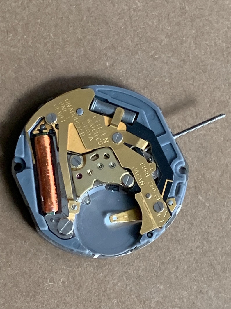 May include: A close-up of a watch movement with gold-colored gears, a copper-colored coil, and a gray plastic base. The text "CITIZEN WATCH CO." and "JAPAN" are visible. A thin metal rod extends from the movement.