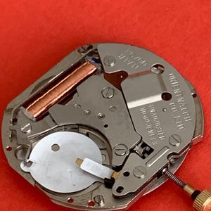 K&ouml;nnte beinhalten: Nahaufnahme eines Uhrwerks mit der Aufschrift "ORIENT WATCH CO., LTD." und "UNADJUSTED NO JEWELS". Das Uhrwerk hat Kupferspulen, silberfarbene Metallkomponenten und eine goldfarbene Schraube. Der Hintergrund ist rot.