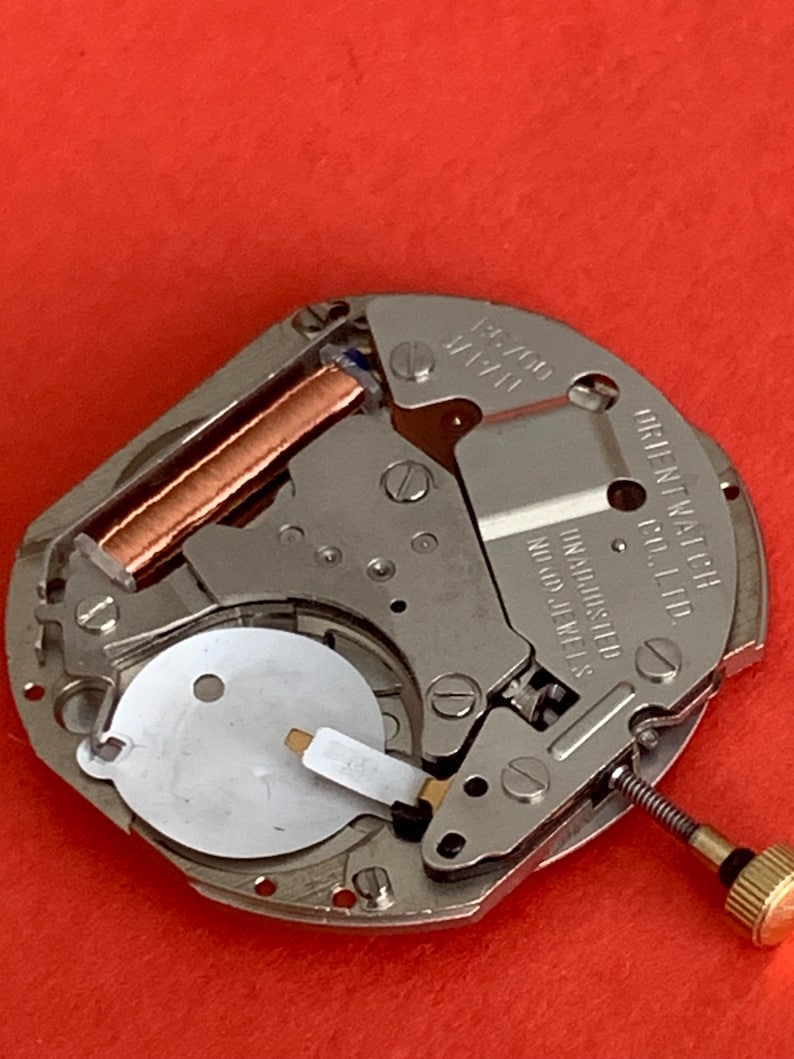 K&ouml;nnte beinhalten: Nahaufnahme eines Uhrwerks mit der Aufschrift "ORIENT WATCH CO., LTD." und "UNADJUSTED NO JEWELS". Das Uhrwerk hat Kupferspulen, silberfarbene Metallkomponenten und eine goldfarbene Schraube. Der Hintergrund ist rot.