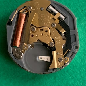 May include: A close-up of a watch movement with gold-colored gears, a copper-colored coil, and a silver-colored battery. The movement is set in a gray plastic casing. The background is a solid green color.