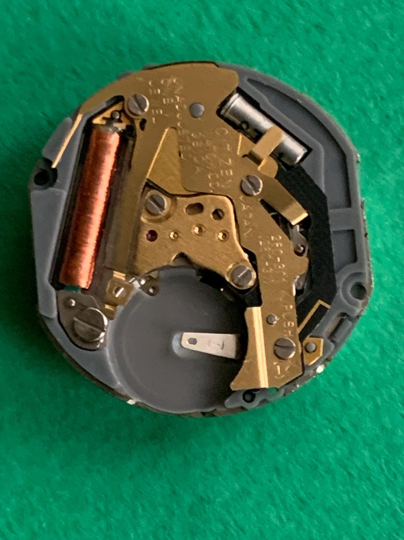 May include: A close-up of a watch movement with gold-colored gears, a copper-colored coil, and a silver-colored battery. The movement is set in a gray plastic casing. The background is a solid green color.
