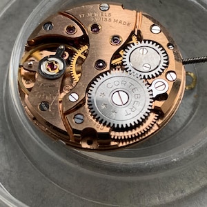 May include: Close-up of a watch movement with visible gears, jewels, and the text "17 JEWELS SWISS MADE." The watch mechanism has a copper-colored base with silver and gold-colored components. The word "CORTEBERT" is visible on a gear.