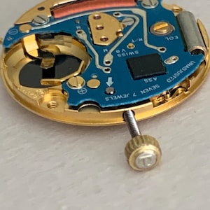 May include: Close-up of a watch movement with a gold-colored frame and a blue circuit board. The board has the text "SWISS V8" and "SEVEN 7 JEWELS". A screw with a gold-colored head is visible.