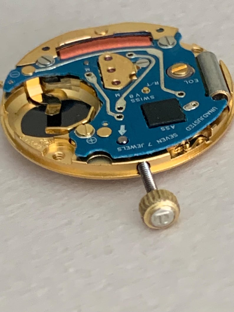 May include: Close-up of a watch movement with a gold-colored frame and a blue circuit board. The board has the text "SWISS V8" and "SEVEN 7 JEWELS". A screw with a gold-colored head is visible.