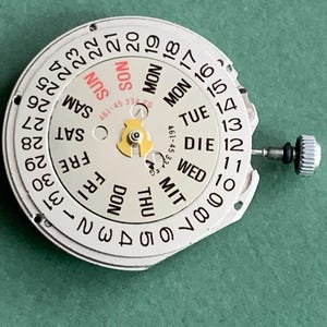 May include: A watch movement with a white dial showing black numbers and day indicators in English, German, and French. The dial displays the days of the week and the date, with a small crown and stem. The text "SUN" is in red.