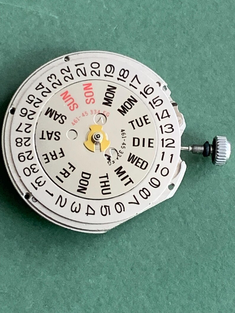 May include: A watch movement with a white dial showing black numbers and day indicators in English, German, and French. The dial displays the days of the week and the date, with a small crown and stem. The text "SUN" is in red.