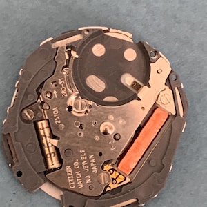 May include: A close-up of a Citizen watch movement, featuring intricate gears, circuits, and a battery. The movement is primarily silver and gray, with a bright orange component and the text "CITIZEN WATCH CO. NJ JEWELS JAPAN".