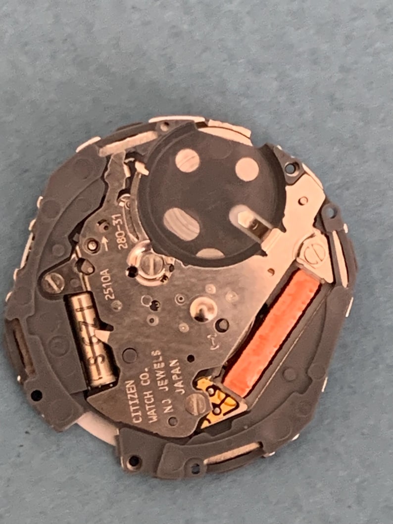 May include: A close-up of a Citizen watch movement, featuring intricate gears, circuits, and a battery. The movement is primarily silver and gray, with a bright orange component and the text "CITIZEN WATCH CO. NJ JEWELS JAPAN".