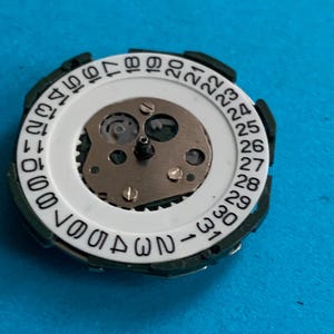 May include: A close-up of a watch movement component. It features a white ring with black numbers from 1 to 31, representing the days of the month. The centre reveals intricate gears and mechanisms, set against a blue background.