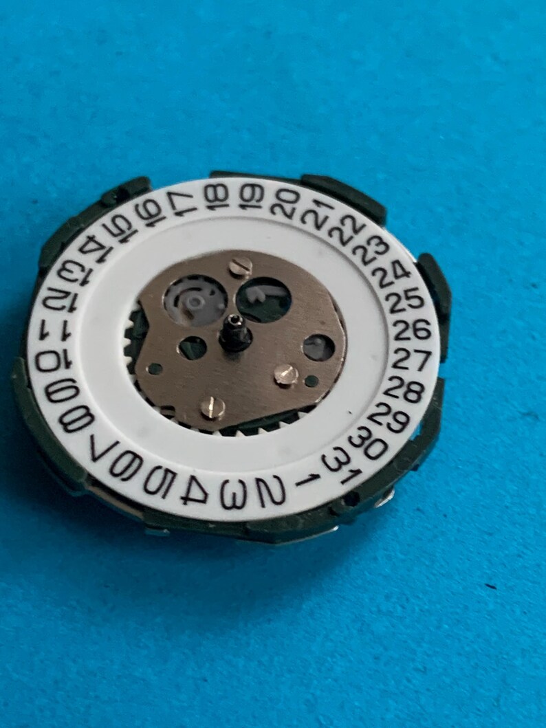 May include: A close-up of a watch movement component. It features a white ring with black numbers from 1 to 31, representing the days of the month. The centre reveals intricate gears and mechanisms, set against a blue background.