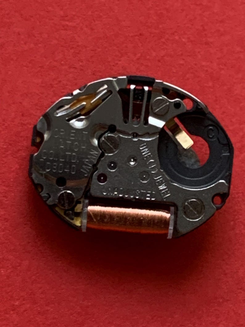 May include: Close-up of a watch movement with silver and gold components. The text "ORIENT WATCH CO., LTD. JAPAN D8040" is visible. The movement includes a copper coil and the text "ONE (1) JEWEL" and "UNADJUSTED".