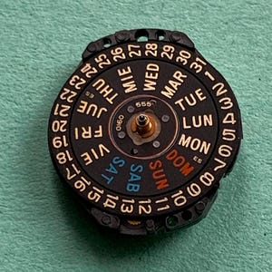 May include: A black watch calendar wheel with white numerals and day abbreviations in multiple languages. The wheel has a central gold-colored post and is set against a green background.