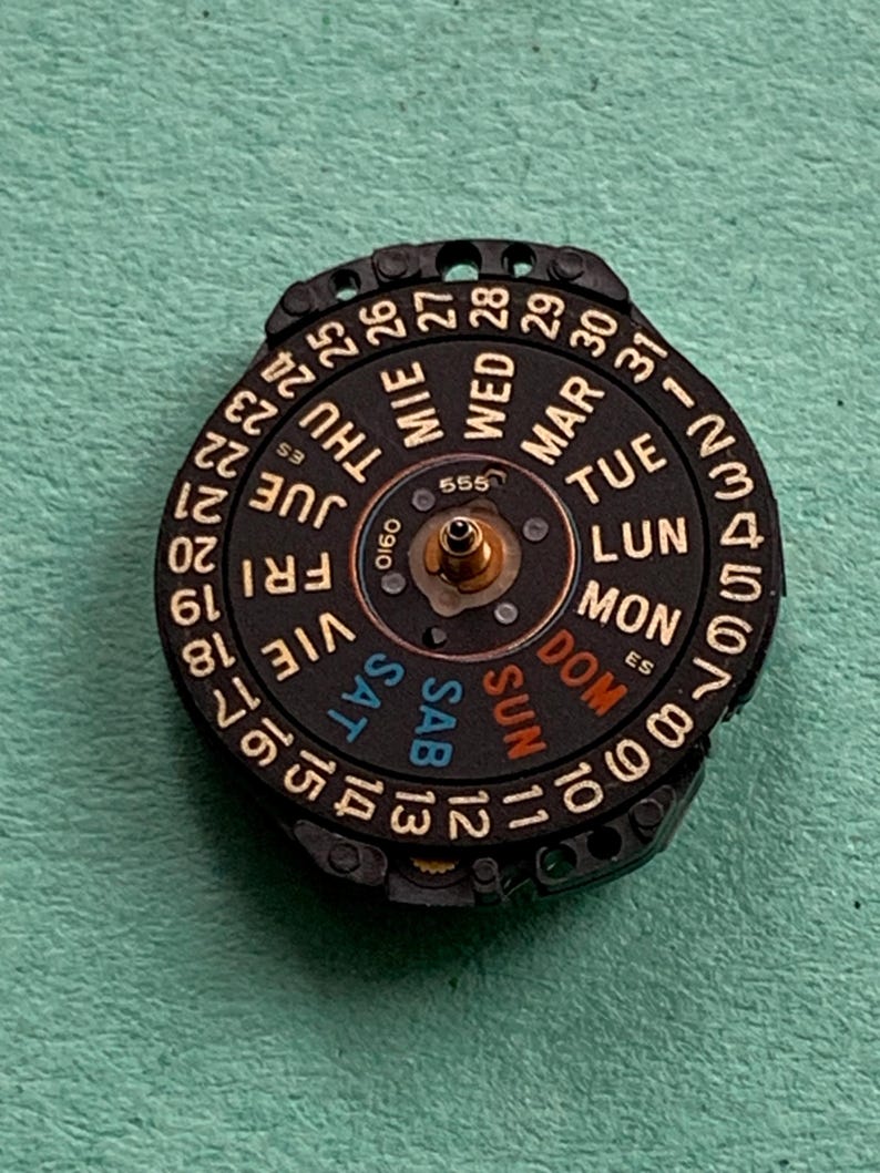 May include: A black watch calendar wheel with white numerals and day abbreviations in multiple languages. The wheel has a central gold-colored post and is set against a green background.