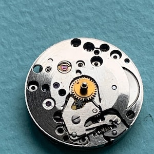 May include: A silver watch movement with a gold gear and a small red jewel. The movement is designed for a wristwatch.