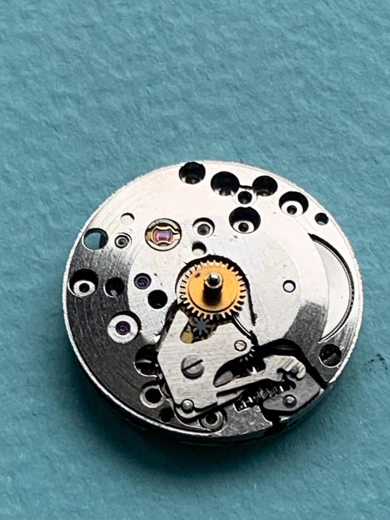 May include: A silver watch movement with a gold gear and a small red jewel. The movement is designed for a wristwatch.