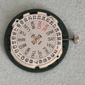 May include: A watch movement with a white dial displaying the days of the week in English and numbers 1-31. The dial is surrounded by a dark green frame and includes a small silver adjustment screw.