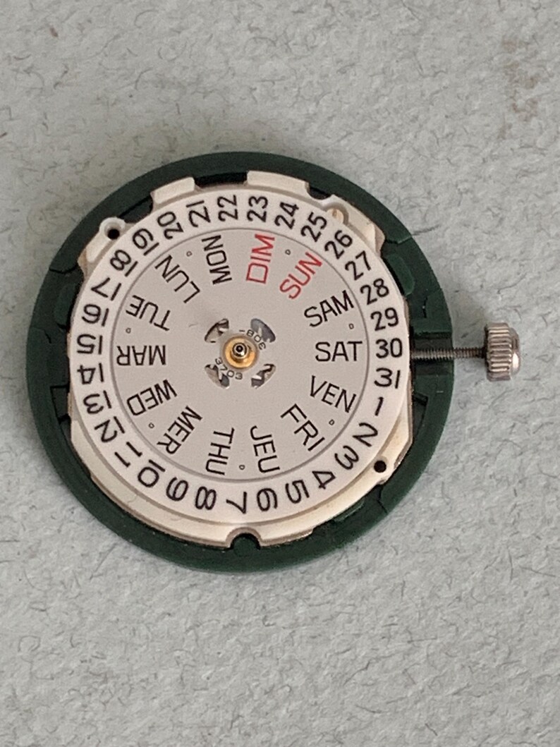 May include: A watch movement with a white dial displaying the days of the week in English and numbers 1-31. The dial is surrounded by a dark green frame and includes a small silver adjustment screw.