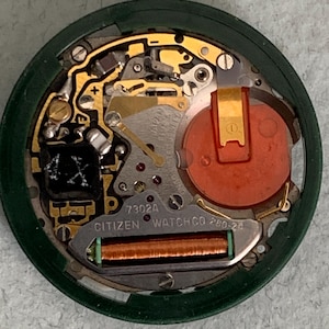May include: Close-up of a Citizen watch movement, showcasing intricate internal components. The watch mechanism features gold-colored gears, a red battery, and the text "CITIZEN WATCH CO". The movement is encased in a green circular frame.