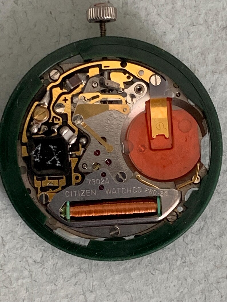 May include: Close-up of a Citizen watch movement, showcasing intricate internal components. The watch mechanism features gold-colored gears, a red battery, and the text "CITIZEN WATCH CO". The movement is encased in a green circular frame.