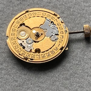 May include: Close-up of a gold-toned watch movement with visible gears, a date dial, and a winding stem. The intricate mechanism displays various components, including small screws and a crown. The background is a solid gray surface.