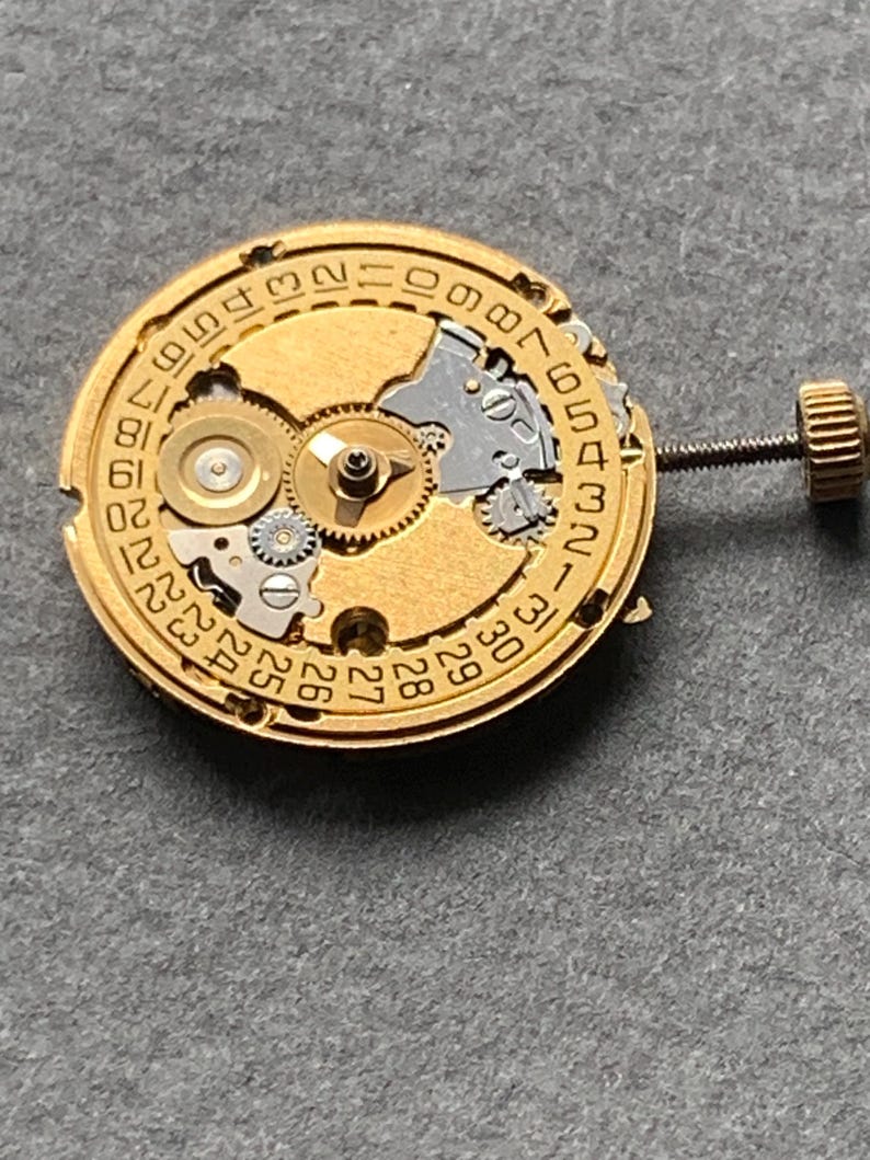 May include: Close-up of a gold-toned watch movement with visible gears, a date dial, and a winding stem. The intricate mechanism displays various components, including small screws and a crown. The background is a solid gray surface.
