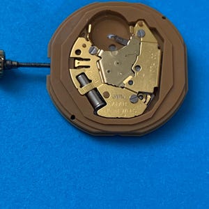 May include: A close-up of a watch movement with gold-colored gears and components set within a brown plastic casing. The watch movement is on a blue surface. The text "16 JEWELS" is visible.