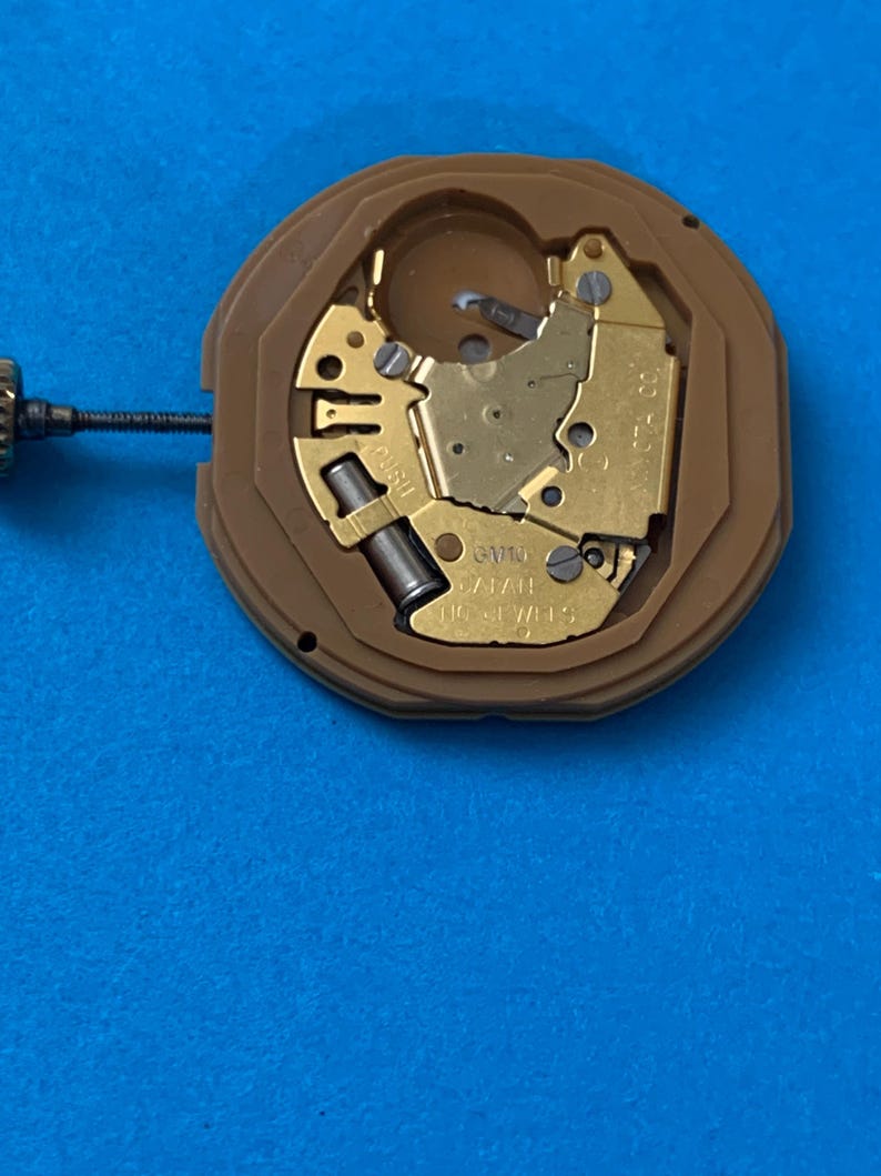 May include: A close-up of a watch movement with gold-colored gears and components set within a brown plastic casing. The watch movement is on a blue surface. The text "16 JEWELS" is visible.