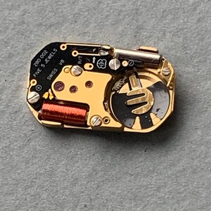 May include: A close-up of a watch movement with gold and black components. The text "280 002 FIVE 5 JEWELS SWISS V8" is visible. The movement has a red coil and various screws and gears.