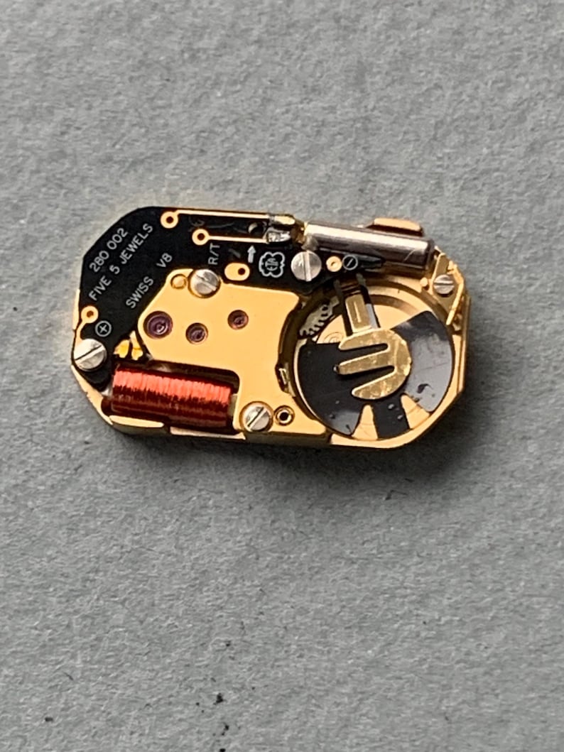 May include: A close-up of a watch movement with gold and black components. The text "280 002 FIVE 5 JEWELS SWISS V8" is visible. The movement has a red coil and various screws and gears.