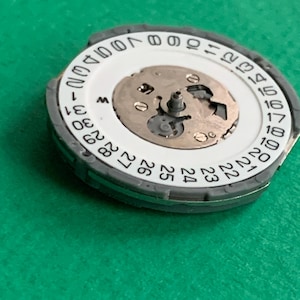 May include: A watch movement with a white date dial and a gray outer ring. The dial displays numbers 1-31, with the day of the week indicator. The central mechanism features small gears and a central post. The background is a solid green.
