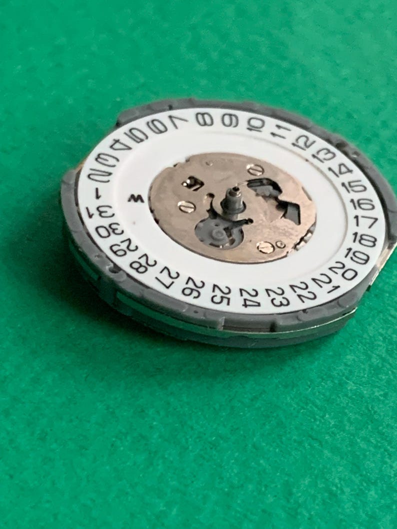 May include: A watch movement with a white date dial and a gray outer ring. The dial displays numbers 1-31, with the day of the week indicator. The central mechanism features small gears and a central post. The background is a solid green.
