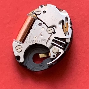 May include: Close-up of a watch movement with intricate gears, screws, and a copper coil. The words "UNADJUSTED" and "ONE (1) JEWEL" are visible. The watch mechanism is set against a vibrant red background.